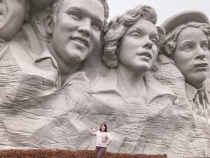 Woman standing in front of attraction that looks like Mt. Rushmore, but has Elvis, Marilyn Monroe, and John Wayne.