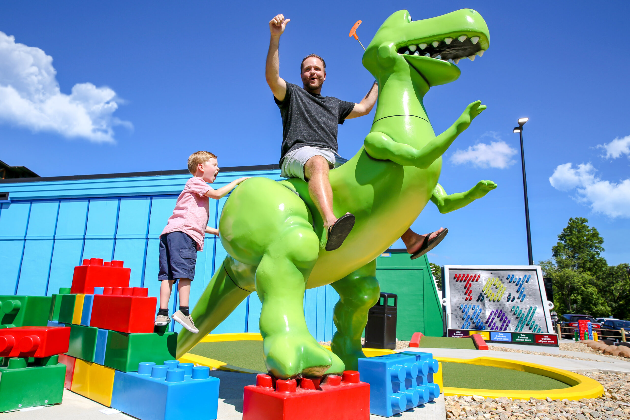 A man on the back of a giant toy dinosaur with his son climbing on the tail.