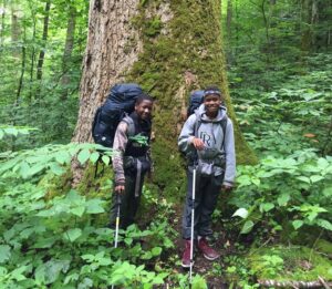 Two young boys standing by a tree smiling and holding walking sticks. 